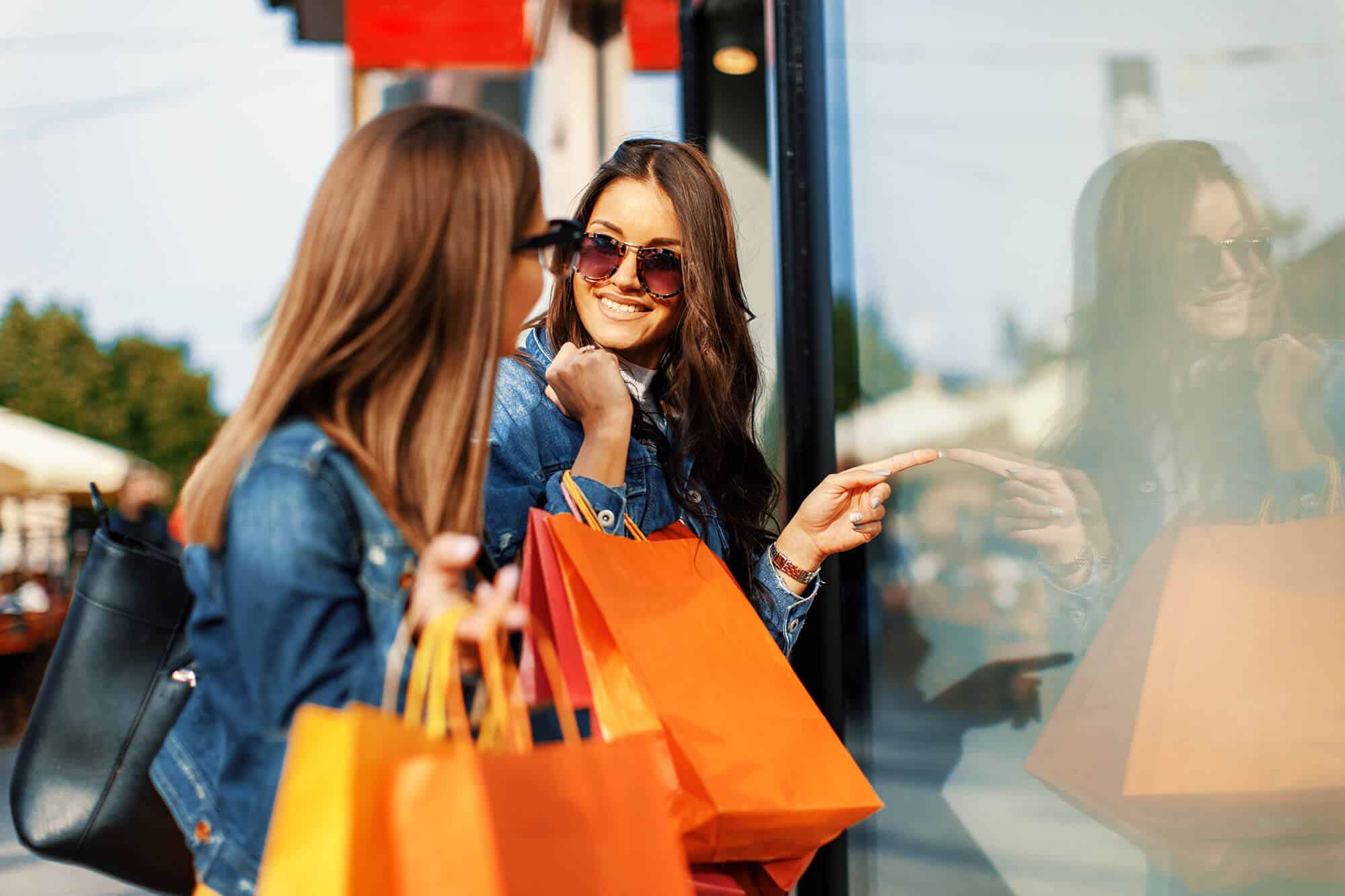 Two girls with shopping bags smiling while pointing in a storefront window